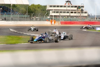 A group of race cars navigating a track, with barriers and a grandstand in the background. The cars are in close competition, moving at high speed with visible motion blur, suggesting a dynamic race environment. Marshals in orange suits are positioned near the track for safety.