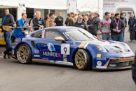 A race car with blue and gold detailing is parked at an event, surrounded by people. The car features multiple sponsor logos and decals, and a British flag is visible on it. Several men stand nearby, engaging with the car or each other. The event appears to be a competitive racing event given the setup and signage.