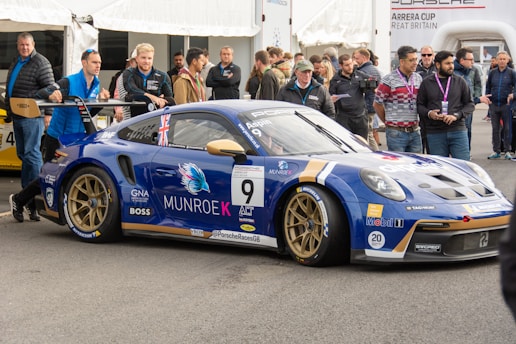 A race car with blue and gold detailing is parked at an event, surrounded by people. The car features multiple sponsor logos and decals, and a British flag is visible on it. Several men stand nearby, engaging with the car or each other. The event appears to be a competitive racing event given the setup and signage.