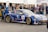A race car with blue and gold detailing is parked at an event, surrounded by people. The car features multiple sponsor logos and decals, and a British flag is visible on it. Several men stand nearby, engaging with the car or each other. The event appears to be a competitive racing event given the setup and signage.