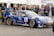 A race car with blue and gold detailing is parked at an event, surrounded by people. The car features multiple sponsor logos and decals, and a British flag is visible on it. Several men stand nearby, engaging with the car or each other. The event appears to be a competitive racing event given the setup and signage.