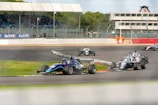 Several race cars are navigating a curve during a motorsport event on a paved track. The cars are open-wheeled, featuring aerodynamic designs, and are competing closely. Spectators can be seen in the stands, and a few marshals in orange suits are present alongside the track near a control tower.