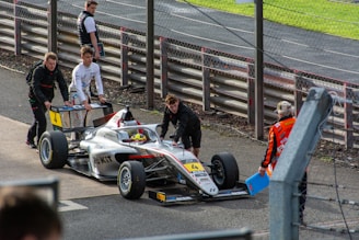 A group of individuals are pushing a racing car along a trackside area. The car is sleek, silver-gray with prominent sponsor logos and the number 4 displayed on its body. Two individuals in racing attire are actively pushing the car, while another person in an orange safety vest supervises. The background features racetrack barriers and fencing, adding to the sporty atmosphere.