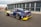 A high-performance race car in a pit area, featuring a sleek design with branding and sponsorship decals. The car is dark blue with a contrasting yellow front bumper and a large rear spoiler. The environment includes a tented pit stop area and a person walking nearby.
