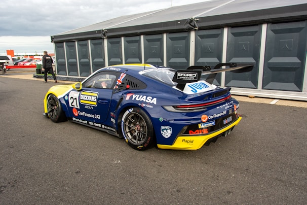 A high-performance race car in a pit area, featuring a sleek design with branding and sponsorship decals. The car is dark blue with a contrasting yellow front bumper and a large rear spoiler. The environment includes a tented pit stop area and a person walking nearby.