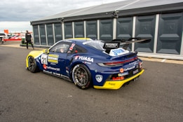 A high-performance race car in a pit area, featuring a sleek design with branding and sponsorship decals. The car is dark blue with a contrasting yellow front bumper and a large rear spoiler. The environment includes a tented pit stop area and a person walking nearby.