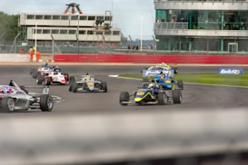 A group of Formula 3 race cars are navigating a racetrack with a few cars leading the pack. In the background, there is a grandstand with spectators, as well as a few marshals standing near the track. The image captures the dynamic motion and competition among the drivers.