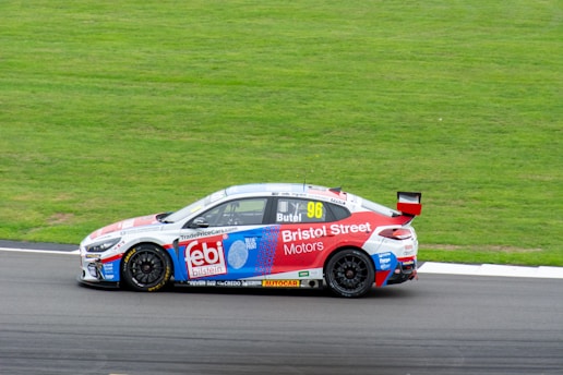 A racing car with the number 96, sponsored by Bristol Street Motors and febi Bilstein, is captured on a race track. The car is moving at high speed with a grassy area and a part of the track visible in the background. The car is predominantly white with red and blue accents and has various sponsor logos on its body.