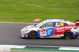 A race car adorned with various sponsor logos speeds along a race track. The car is predominantly red, white, and blue, with the number 96 prominently displayed on its side. The surroundings include a well-kept asphalt track and green grass, suggesting it is on a motor racing circuit.