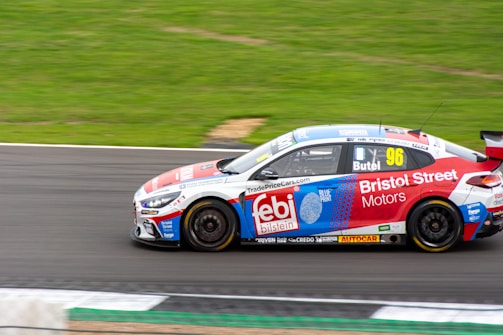 A race car adorned with various sponsor logos speeds along a race track. The car is predominantly red, white, and blue, with the number 96 prominently displayed on its side. The surroundings include a well-kept asphalt track and green grass, suggesting it is on a motor racing circuit.