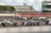 A group of race cars navigating a track during a motorsport event, with drivers intensely focused as they maneuver through a curve. The setting includes a large trackside building with spectators observing the race. Marshals in orange suits stand by, ensuring safety. Advertising banners and racing numbers are visible on the cars and track.