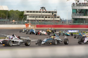 A group of race cars navigating a track during a motorsport event, with drivers intensely focused as they maneuver through a curve. The setting includes a large trackside building with spectators observing the race. Marshals in orange suits stand by, ensuring safety. Advertising banners and racing numbers are visible on the cars and track.