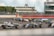 A group of race cars navigating a track during a motorsport event, with drivers intensely focused as they maneuver through a curve. The setting includes a large trackside building with spectators observing the race. Marshals in orange suits stand by, ensuring safety. Advertising banners and racing numbers are visible on the cars and track.