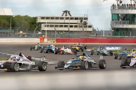A group of race cars navigating a track during a motorsport event, with drivers intensely focused as they maneuver through a curve. The setting includes a large trackside building with spectators observing the race. Marshals in orange suits stand by, ensuring safety. Advertising banners and racing numbers are visible on the cars and track.