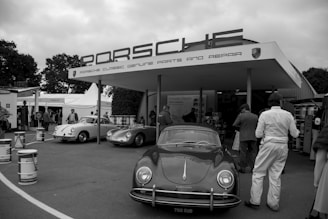 A vintage car exhibit with several classic Porsche vehicles displayed outdoors. A large sign reading 'Porsche Classic Genuine Parts and Repair' is visible above the stand, where several people are gathered. The setting has a retro vibe, with barrels and banners complementing the historical theme. Trees and tents are seen in the background, adding to the exhibition atmosphere.