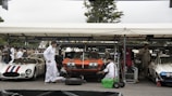 A parking area with classic cars where mechanics dressed in white overalls are working on vehicles. The cars include a red and white striped Maserati, an orange Chevrolet Corvette, and a white Corvette, all parked under a marquee. Several people are gathered around, and the environment suggests a vintage car event.