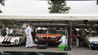 A parking area with classic cars where mechanics dressed in white overalls are working on vehicles. The cars include a red and white striped Maserati, an orange Chevrolet Corvette, and a white Corvette, all parked under a marquee. Several people are gathered around, and the environment suggests a vintage car event.