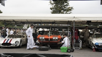 A parking area with classic cars where mechanics dressed in white overalls are working on vehicles. The cars include a red and white striped Maserati, an orange Chevrolet Corvette, and a white Corvette, all parked under a marquee. Several people are gathered around, and the environment suggests a vintage car event.