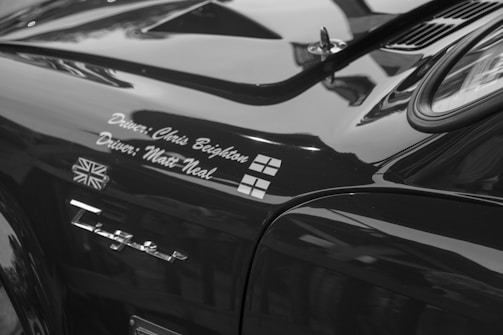 A close-up view of a car with reflective black paint, featuring the names of two drivers written in cursive script. There are two small flags, the Union Jack and another with a cross, displayed near the names. The word 'E-Type' is prominently shown as a badge below the names.