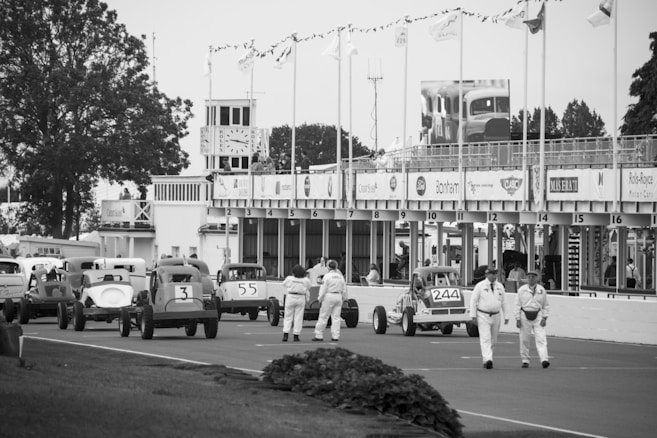 Vintage race cars are lined up on a racetrack, with a group of people dressed in period clothing walking nearby. The scene includes a building with multiple flags and a large clock, alongside trees and classic advertisements. The atmosphere suggests an old-fashioned or retro racing event.