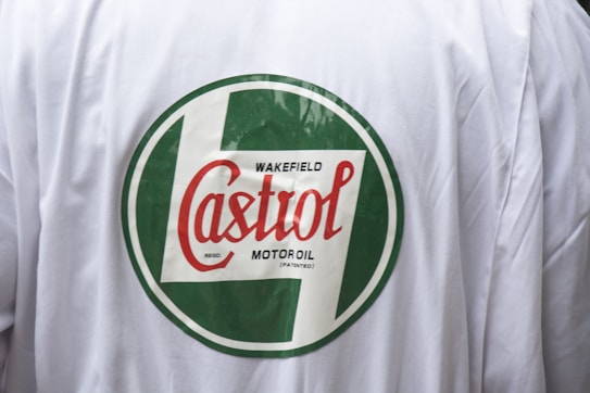 A close-up view of a white jacket with a large, circular logo in green and red. The logo prominently features the name 'Castrol', along with additional text 'Wakefield' and 'Motor Oil'. The text is stylishly arranged within the circle, and the backdrop is a plain white fabric.