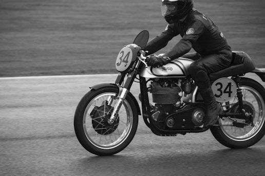 A motorcyclist wearing protective gear is riding a vintage Norton motorcycle on a racetrack. The motorcycle displays the number 34 on both the front plate and the side plate, and the image is captured in black and white.