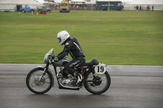 A motorcyclist dressed in black leather gear and a white helmet rides a vintage motorcycle on a racetrack. The motorcycle is marked with the number 19, and the backdrop is a green grassy area with tents and vehicles in the distance.