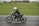 A motorcyclist dressed in black leather gear and a white helmet rides a vintage motorcycle on a racetrack. The motorcycle is marked with the number 19, and the backdrop is a green grassy area with tents and vehicles in the distance.
