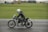 A motorcyclist dressed in black leather gear and a white helmet rides a vintage motorcycle on a racetrack. The motorcycle is marked with the number 19, and the backdrop is a green grassy area with tents and vehicles in the distance.