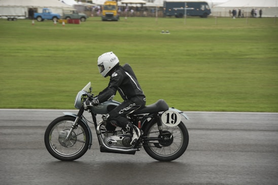 A motorcyclist dressed in black leather gear and a white helmet rides a vintage motorcycle on a racetrack. The motorcycle is marked with the number 19, and the backdrop is a green grassy area with tents and vehicles in the distance.