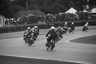 A group of motorcyclists are racing on a track, viewed from behind as they lean into a curve. The riders are wearing helmets and racing gear. Spectators with umbrellas are visible behind a fence, indicating cloudy or rainy weather. The setting has a vintage feel, and the image is in black and white.