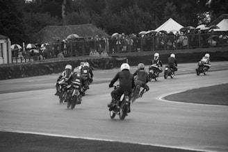 A group of motorcyclists are racing on a track, viewed from behind as they lean into a curve. The riders are wearing helmets and racing gear. Spectators with umbrellas are visible behind a fence, indicating cloudy or rainy weather. The setting has a vintage feel, and the image is in black and white.