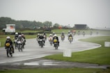 A group of motorcyclists dressed in racing gear is riding vintage motorcycles on a wet racetrack. The track is surrounded by green grass and a few spectators can be seen in the distance under a cloudy sky. The scene suggests a competitive race despite the rainy conditions.