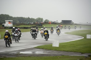 A group of motorcyclists dressed in racing gear is riding vintage motorcycles on a wet racetrack. The track is surrounded by green grass and a few spectators can be seen in the distance under a cloudy sky. The scene suggests a competitive race despite the rainy conditions.