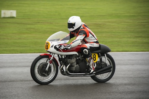 A motorcyclist in racing gear, including a helmet and a protective suit, is riding a race bike on a wet track. The bike has a number 78 displayed on the front and side, and there is a blurred motion suggesting speed. The background is a grassy area, indicating an outdoor race circuit.