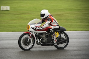 A motorcyclist in racing gear, including a helmet and a protective suit, is riding a race bike on a wet track. The bike has a number 78 displayed on the front and side, and there is a blurred motion suggesting speed. The background is a grassy area, indicating an outdoor race circuit.