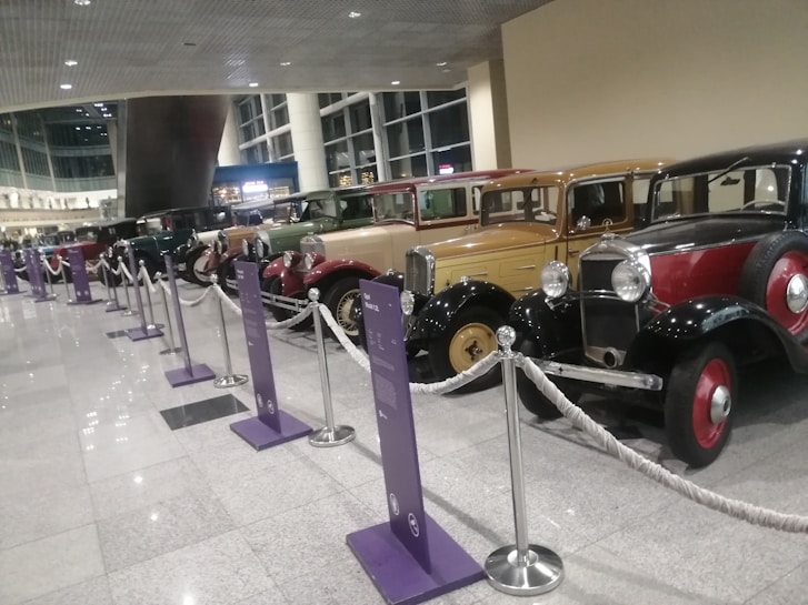 A row of vintage cars displayed in a well-lit indoor exhibition area, separated by ropes and informational signs on stands. The cars vary in color and style, reflecting different eras of automotive design, and are positioned on a glossy tiled floor. The background features large windows and architectural details, creating a modern contrast to the classic vehicles.