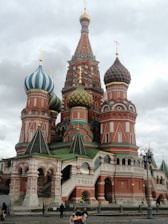An ornate building with colorful, onion-shaped domes featuring intricate patterns and bright hues of red, green, and blue. The structure includes detailed arches and spires, showcasing classic Russian architecture. Several people are seen near the base of the building.