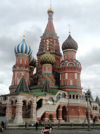 An ornate building with colorful, onion-shaped domes featuring intricate patterns and bright hues of red, green, and blue. The structure includes detailed arches and spires, showcasing classic Russian architecture. Several people are seen near the base of the building.