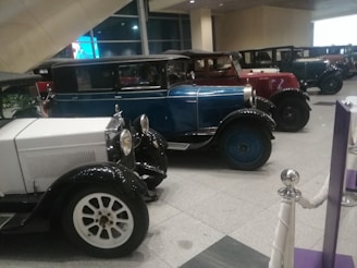 A collection of vintage cars is lined up indoors. The vehicles are parked in an organized row, showcasing various models with distinct designs. The colors include blue, red, and white. The setting appears to be a museum or showroom with smooth tiled flooring and barriers indicating a display area.