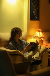 Person wearing trendy eyeglasses smiling while reading a book in a cozy room