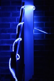 A series of blue LED lights are wrapped around the railing of a staircase against a backdrop of brick walls. The lights create a glowing effect in the dark environment, highlighting the railing and casting a soft blue hue.