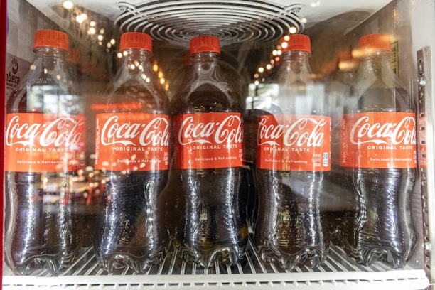 Five Coca-Cola bottles are lined up inside a refrigerated display case. Each bottle has a red label and cap, with condensation visible on the plastic. The bottle labels read 'Coca-Cola Original Taste' along with slogans like 'Delicious & Refreshing'.