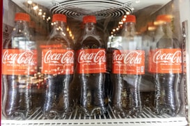 Five Coca-Cola bottles are lined up inside a refrigerated display case. Each bottle has a red label and cap, with condensation visible on the plastic. The bottle labels read 'Coca-Cola Original Taste' along with slogans like 'Delicious & Refreshing'.