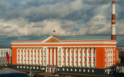 A large neoclassical building with a red and white facade, featuring numerous columns and decorative golden accents. A Russian flag is visible on top of the building, and there is a tall cylindrical chimney with the number 1985 on it in the background. The image captures the building under a cloudy sky, with parking spaces and streets surrounding it.