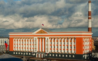 A large neoclassical building with a red and white facade, featuring numerous columns and decorative golden accents. A Russian flag is visible on top of the building, and there is a tall cylindrical chimney with the number 1985 on it in the background. The image captures the building under a cloudy sky, with parking spaces and streets surrounding it.