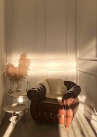 An elegant room features a brown leather armchair with a plush white fur draped over it, situated next to a set of minimalist nesting tables. Delicate, feathery pampas grass is arranged in vases beside the armchair. The room is bathed in warm, natural light streaming through the window, casting gentle shadows on the wall, which has a classic panel design.