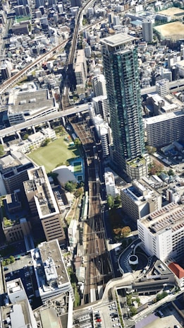 Aerial view of a densely populated urban area featuring a tall modern skyscraper, several mid-rise buildings, and a network of railway tracks that pass through the city. The landscape includes a mix of residential and commercial structures with roads and green spaces interspersed throughout. The architecture reveals a vibrant and bustling urban life.
