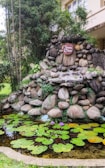 A serene stone fountain surrounded by lush greenery with a sign saying 'Go Green, Breath Clean.' The foreground features a small pond with green lily pads and vibrant purple water lilies.