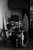 A black drum set is positioned in an indoor setting, surrounded by surfboards. The drums display a logo with the letters 'TH', and the surrounding area appears decorated with textured walls and plants, suggesting a casual or beach-themed ambiance.